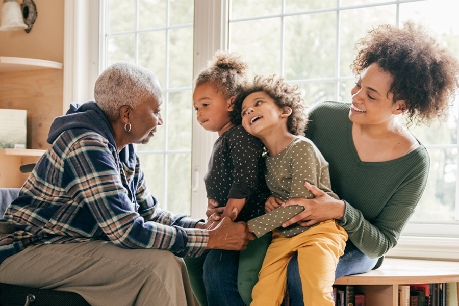 multigenerational family at home. Black grandmother in wheelchair talking with two toddler granddaughters who are sitting on their mother's lap. Sandwich generation.