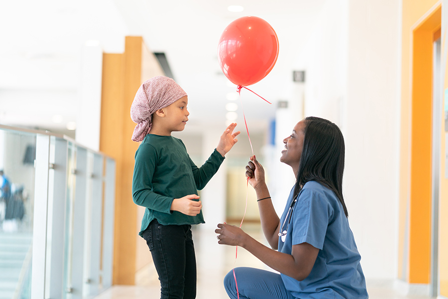 female doctor kneeling in hospital hallway and handing a red balloon to a young cancer patient. demonstrating the importance of empathy in healthcare.