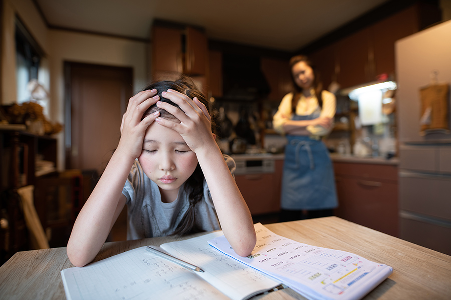 signs of anxiety in children. anxious child 8-10 years old sitting at kitchen table with her head in her hands, elbows on math homework.