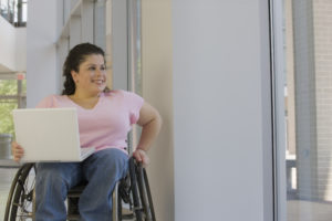 smiling adult woman with spina bifiida, sitting in a wheelchair inside a professional building with an open laptop on her lap. Exploring health issues in adults with Spina Bifida