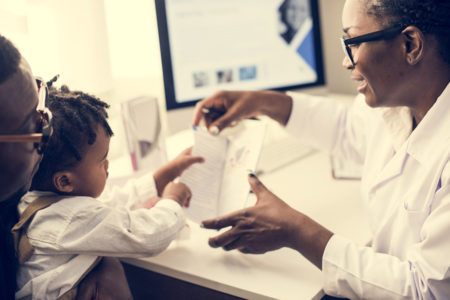 smiling mature black doctor sitting in her office with a parent and young child, showing them something on her desk and explaining what Medicaid Waivers are.