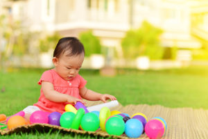 young asian child with hidden feeding tube sitting outside on a blanket int heh grass, looking down with multiple colored balls around her. her family is adjusting to tube feeding a child at home.