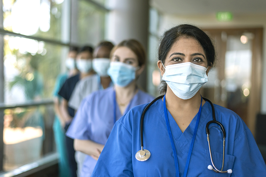 What is a WOC nurse? Row of doctors and nurses standing inside a hospital hallway or lobby, with a smiling, masked female nurse of color at the front.