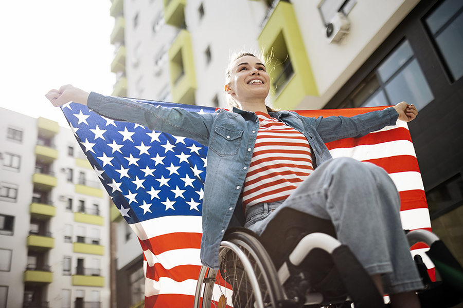 smiling woman in a wheelchair on the streets of a city, holding the American flag spread behind her shoulders. Celebrating National Disability Independence Day.