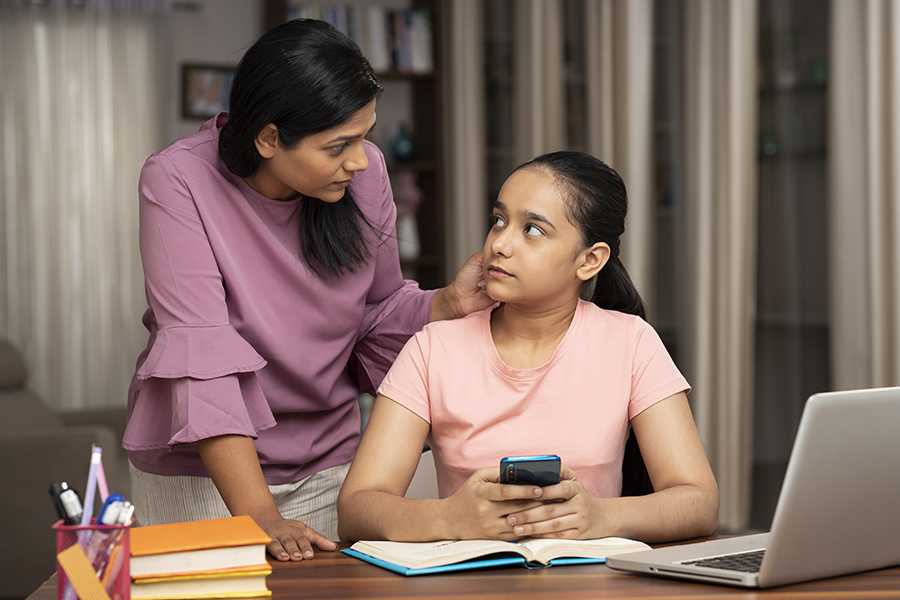 mother leans over and gently confronts teen daughter who is sitting in front of her school work playing on her phone. how to stop being the people pleasing parent.