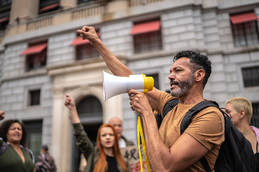 image of man leading strike outside of a multistory building, holding a megaphone with his hand in the air. UPS and Teamsters reach tentative agreement, potentially averting strike