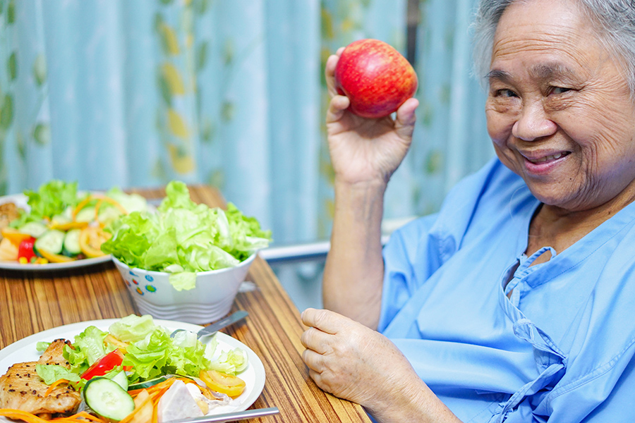 image of older woman smiling before a plateful of healthy food. Woman is wearing hospital scrubs. Malnutrition Awareness Week.